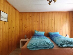 A room with wooden panel walls, a bed with blue bedding, a nightstand with a lamp, and a ceiling fan. - Schwendli Appartement/Fewo, Dusche, WC 3-Zimmerwohnung by Interhome (Kandersteg)