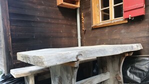 A rustic wooden bench with a bench seat and a table top, positioned outdoors against a wooden wall with a window and a red shutter. - Schwendli Appartement/Fewo, Dusche, WC 3-Zimmerwohnung by Interhome (Kandersteg)