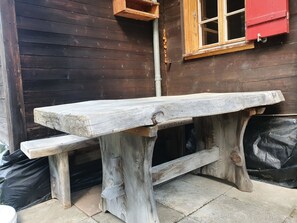 A rustic wooden bench with a bench seat and a table top, positioned outdoors against a wooden wall with a window and a red shutter. - Schwendli Appartement/Fewo, Dusche, WC 3-Zimmerwohnung by Interhome (Kandersteg)