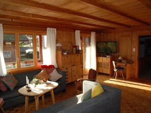 Ein gemütliches Blockhaus-Interieur mit Holzbalkendecke, einem Fernseher, einer Couch, einem Esstisch und einer Küchenzeile.