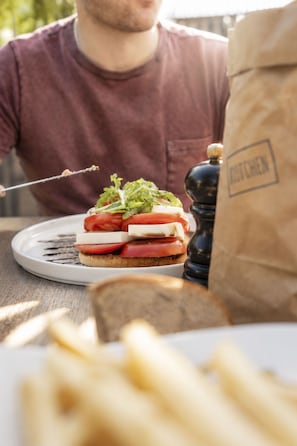 A sandwich on a plate with a person in the background, a pepper grinder, and a paper bag. - Schermtanne Appartement/Fewo, Bad, WC, 1 Schlafraum Wohnung Nr 4 by Interhome (Adelboden)