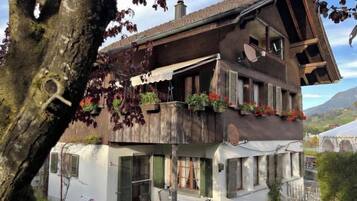Un bâtiment à deux étages doté d’un balcon, de fenêtres avec volets et d’un arbre au premier plan.