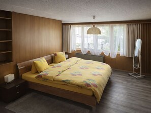Ein Schlafzimmer mit einem Holzbett, einem Nachttisch, einem Heizlüfter und einem Fenster mit Vorhängen.
