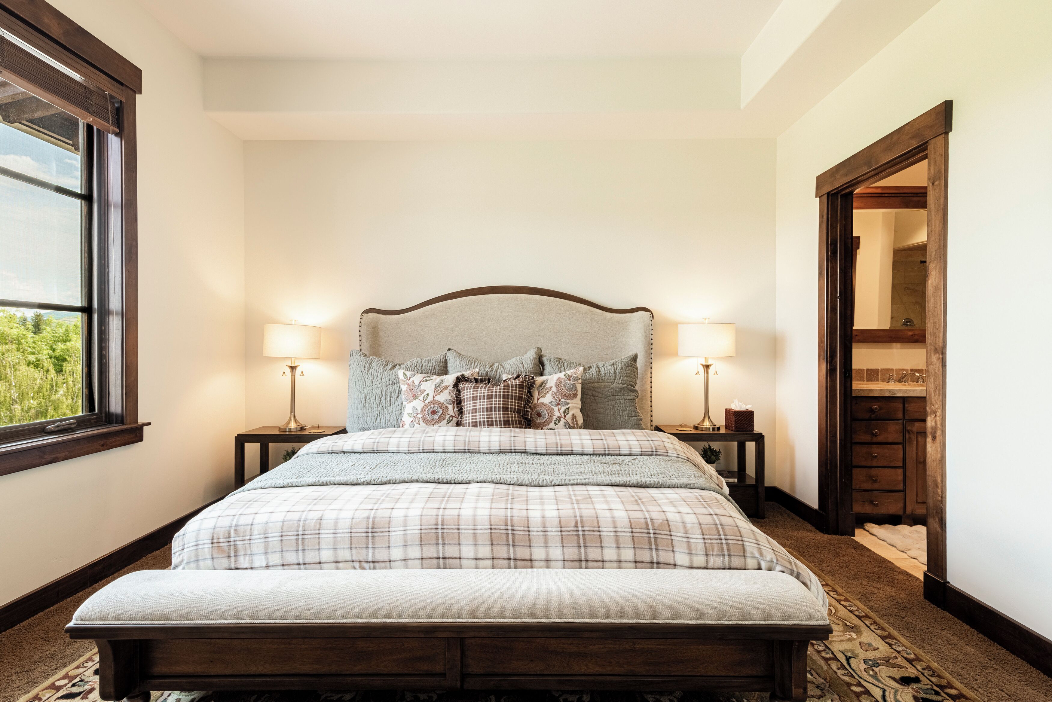 A neatly made bed with a headboard, two bedside lamps, and a view of a room with a wooden door and a window.