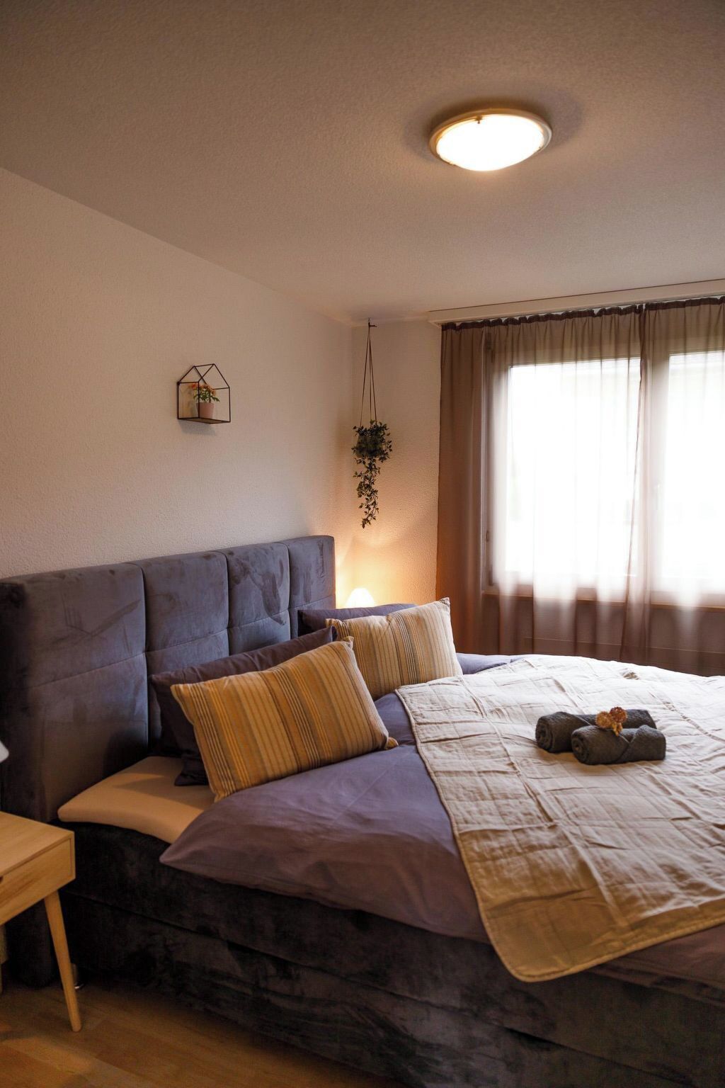 Un dormitorio con cama, mesita de noche, planta colgante y ventana con cortinas.