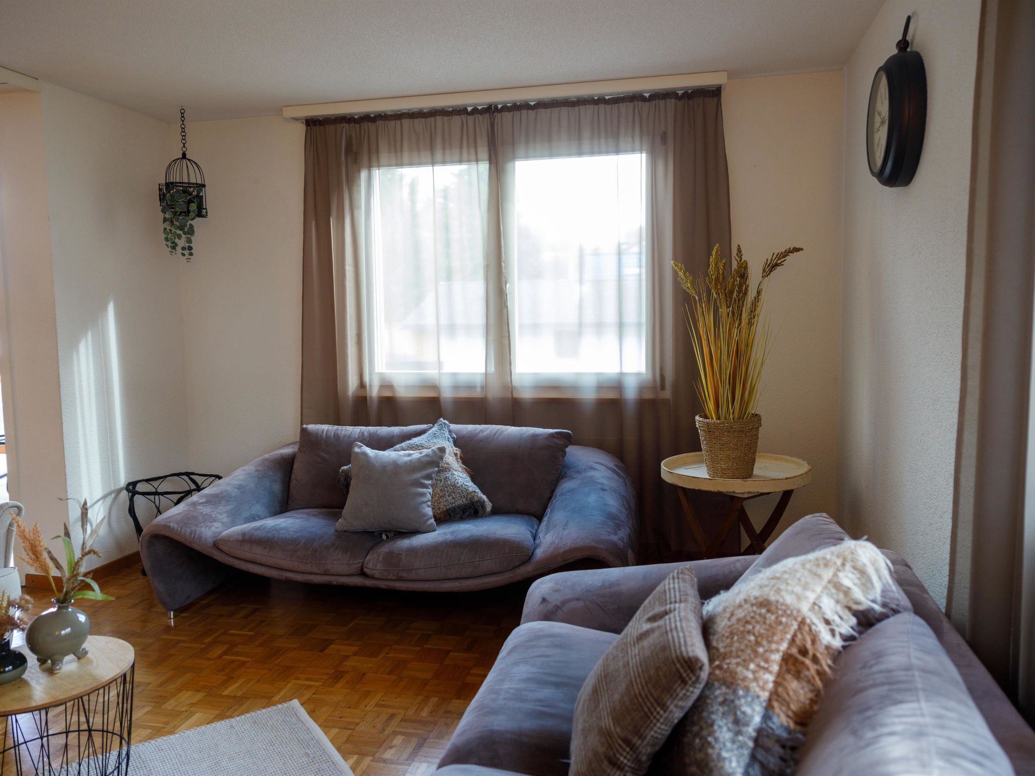 Een woonkamer met een bank, een zithoekje, een tafeltje met een plant en een klok aan de muur.