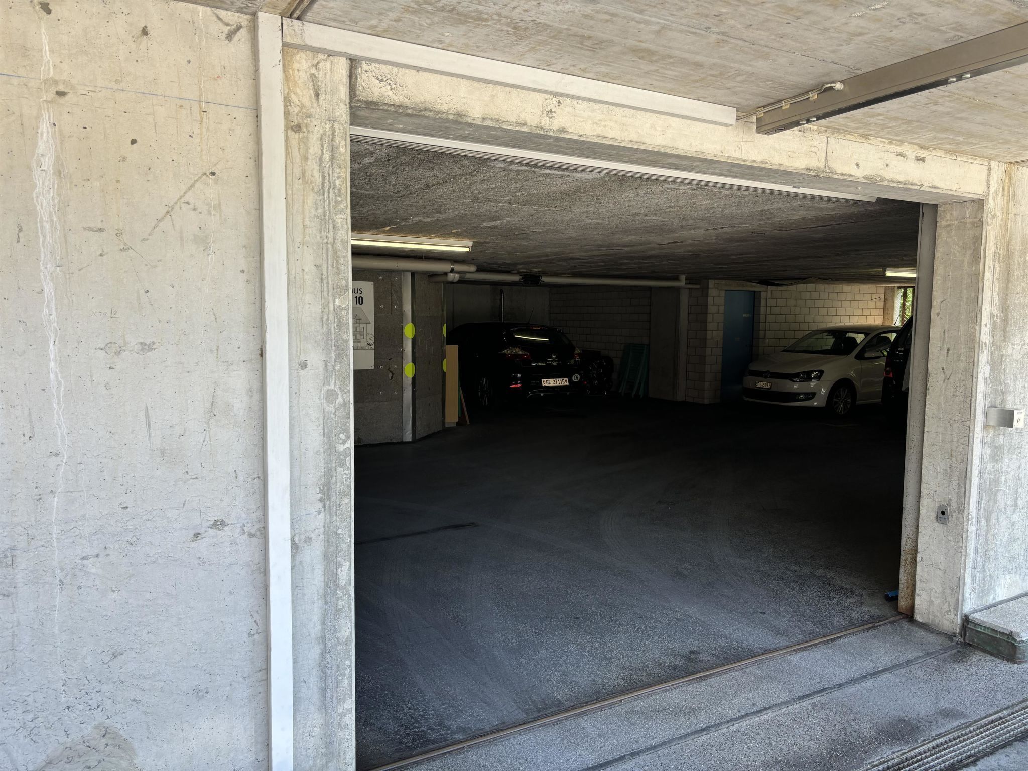 Un parking souterrain aux murs en béton, avec quelques voitures garées et une porte de sortie bien visible.