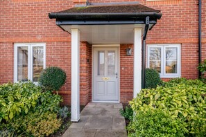 A brick house with a white door, a covered porch, and a small garden with bushes. - Oxfordshire home Surrounded by Greenery (Bessels Leigh)