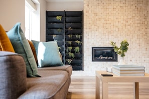 A living room with a sofa, cushions, a coffee table, a black shelving unit with plants, a tiled wall, and a framed picture. - Oxfordshire home Surrounded by Greenery (Bessels Leigh)