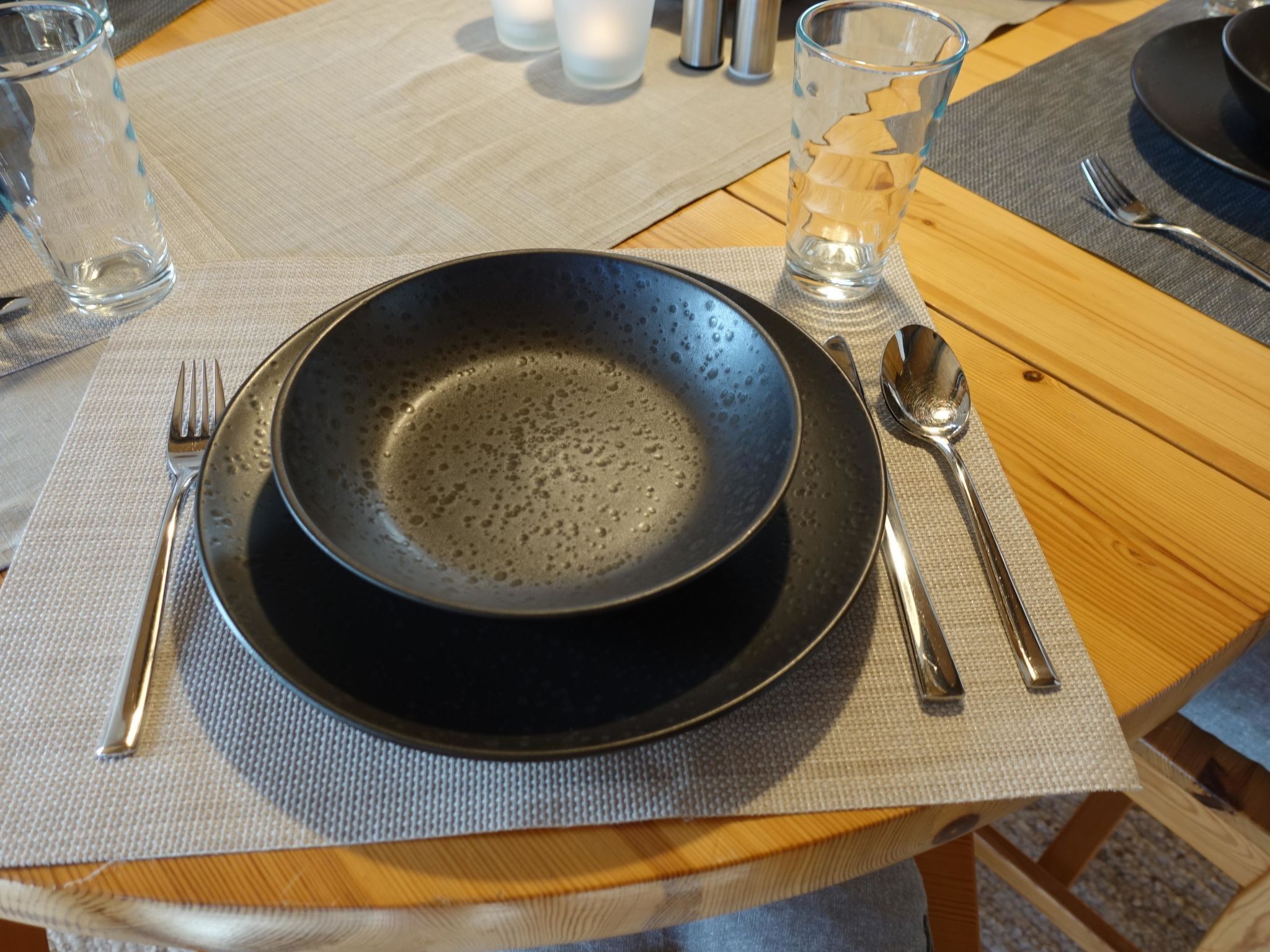 Ein gedeckter Tisch mit schwarzem Teller, Gabel, Löffel und einem Glas Wasser.