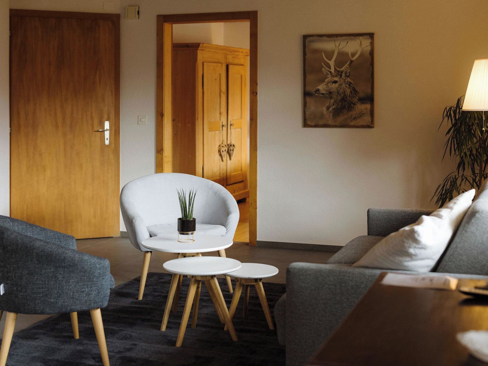 Un salon avec un canapé gris, un fauteuil blanc et une petite table ronde sur laquelle est posée une plante. On y trouve une porte en bois et un tableau encadré accroché au mur.