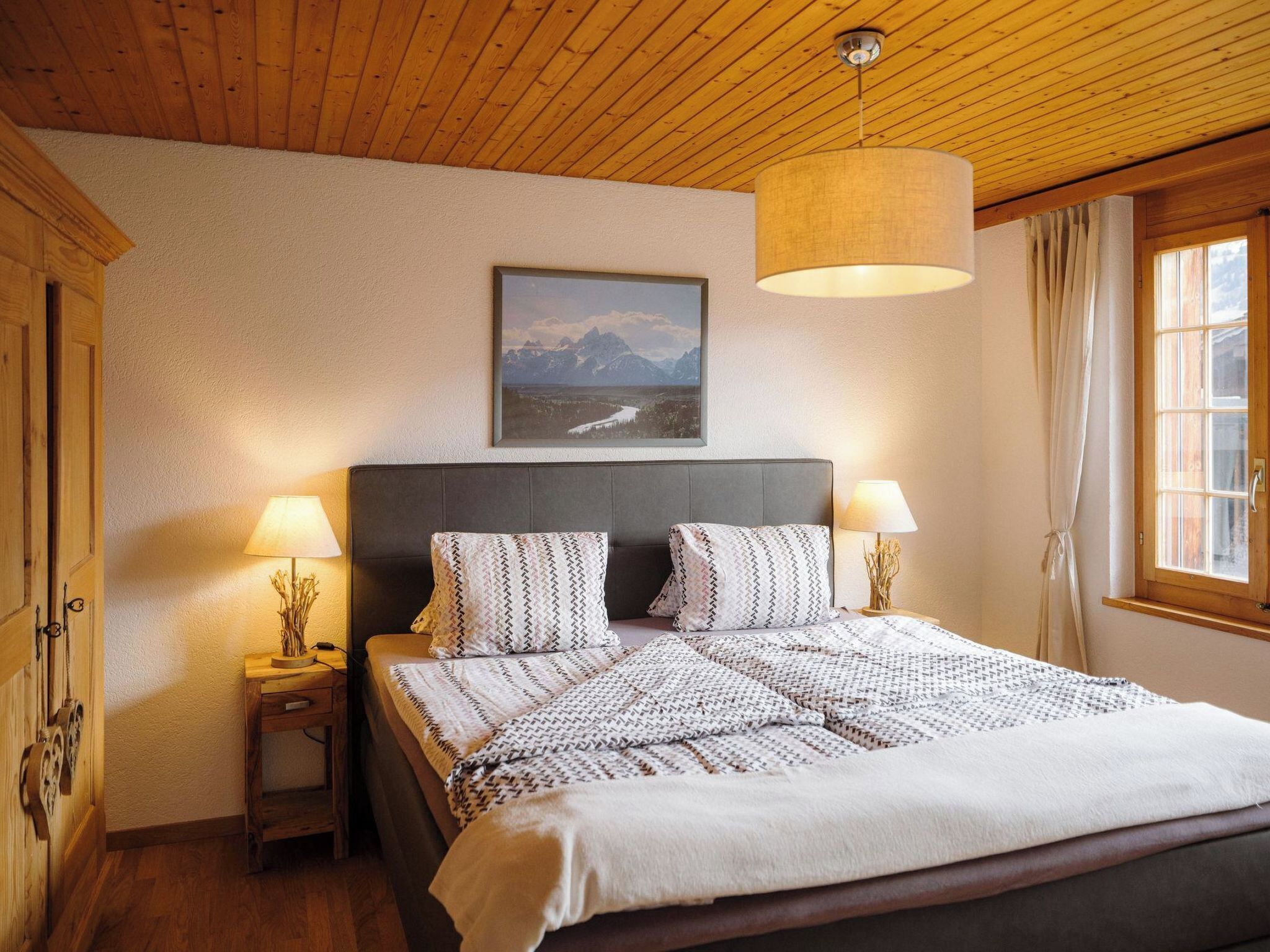 Ein Schlafzimmer mit einem Bett, zwei Nachttischlampen, einem Nachttisch, einem Holzkopfende, einem gerahmten Bild an der Wand und einem Fenster mit Vorhängen.