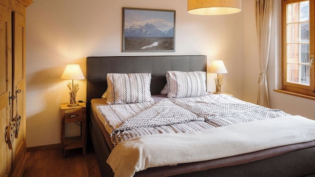 Een slaapkamer met een bed, twee bedlampen, een nachtkastje, een houten hoofdeinde, een ingelijste foto aan de muur en een raam met gordijnen.