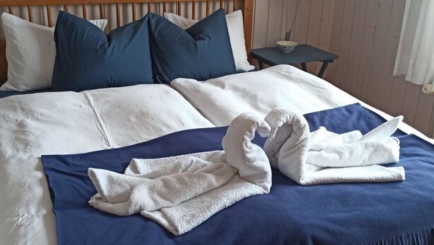 A neatly made bed with a blue bedspread and white pillows, a wooden headboard, a small bedside table with a lamp, and a wall-mounted wreath. - Chalet Edelweiss Appartement/Fewo, Dusche, WC, 2 Schlafräume 3-Zimmerwohnung by Interhome (Frutigen)