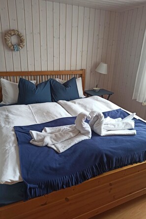A neatly made bed with a blue bedspread and white pillows, a wooden headboard, a small bedside table with a lamp, and a wall-mounted wreath. - Chalet Edelweiss Appartement/Fewo, Dusche, WC, 2 Schlafräume 3-Zimmerwohnung by Interhome (Frutigen)