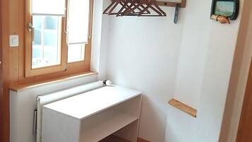 Une pièce avec un bureau blanc, une fenêtre avec des stores, un portant à vêtements et un radiateur.