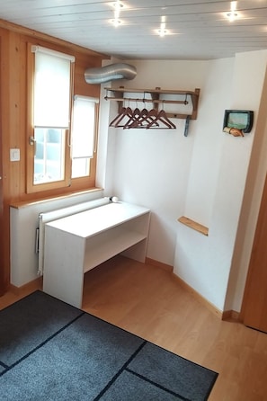 Una habitació amb escriptori blanc, finestra amb persianes, estant penjant per a roba i radiador.