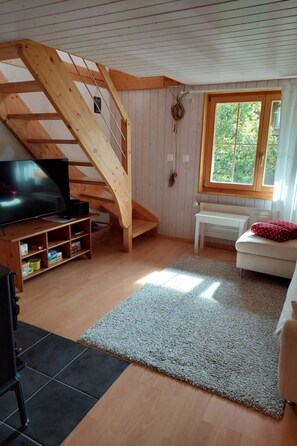 A room with a wooden staircase, a television on a wooden stand, a white sofa, and a window with a view of greenery. - Chalet Edelweiss Appartement/Fewo, Dusche, WC, 2 Schlafräume 3-Zimmerwohnung by Interhome (Frutigen)