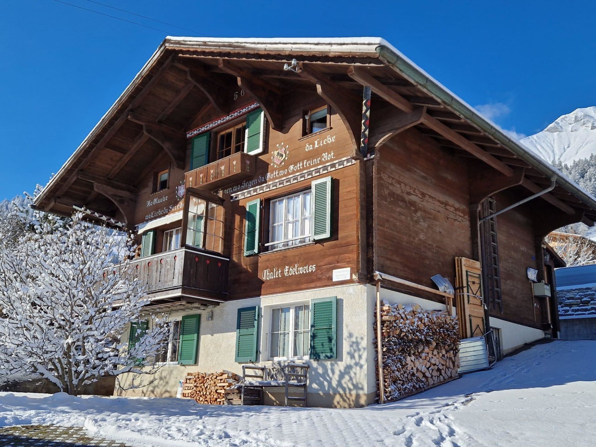 Un chalet en bois doté d’un balcon, une pancarte sur laquelle on peut lire « Da Liebe Gott macht schöne Dinge », et un paysage montagneux en arrière-plan.