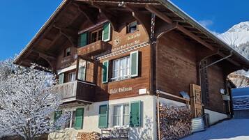 Un chalet en bois doté d’un balcon, une pancarte sur laquelle on peut lire « Da Liebe Gott macht schöne Dinge », et un paysage montagneux en arrière-plan.