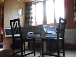 A room with a wooden interior, a table with a blue tablecloth, two chairs, a window with curtains, and a radiator. - Gehret Appartement/Fewo, Dusche, WC, 1 Schlafraum 2-Zimmerwohnung by Interhome (Adelboden)