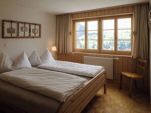 A room with two beds, a wooden chair, a window with curtains, and a radiator. - Gästehaus Alpengruss Ferienhaus, Dusche, WC, 1 Schlafraum Chalet by Interhome (Adelboden)