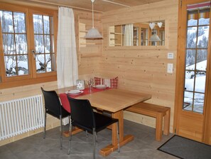 A wooden interior with a dining table, chairs, and a bench. There are windows with curtains, a hanging light fixture, and a view of a snowy landscape. - Gästehaus Alpengruss Ferienhaus, Dusche, WC, 1 Schlafraum Chalet by Interhome (Adelboden)
