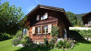 Un chalet en bois doté d’un toit à pente, de grandes fenêtres et d’un jardin bien entretenu où poussent diverses plantes et fleurs.