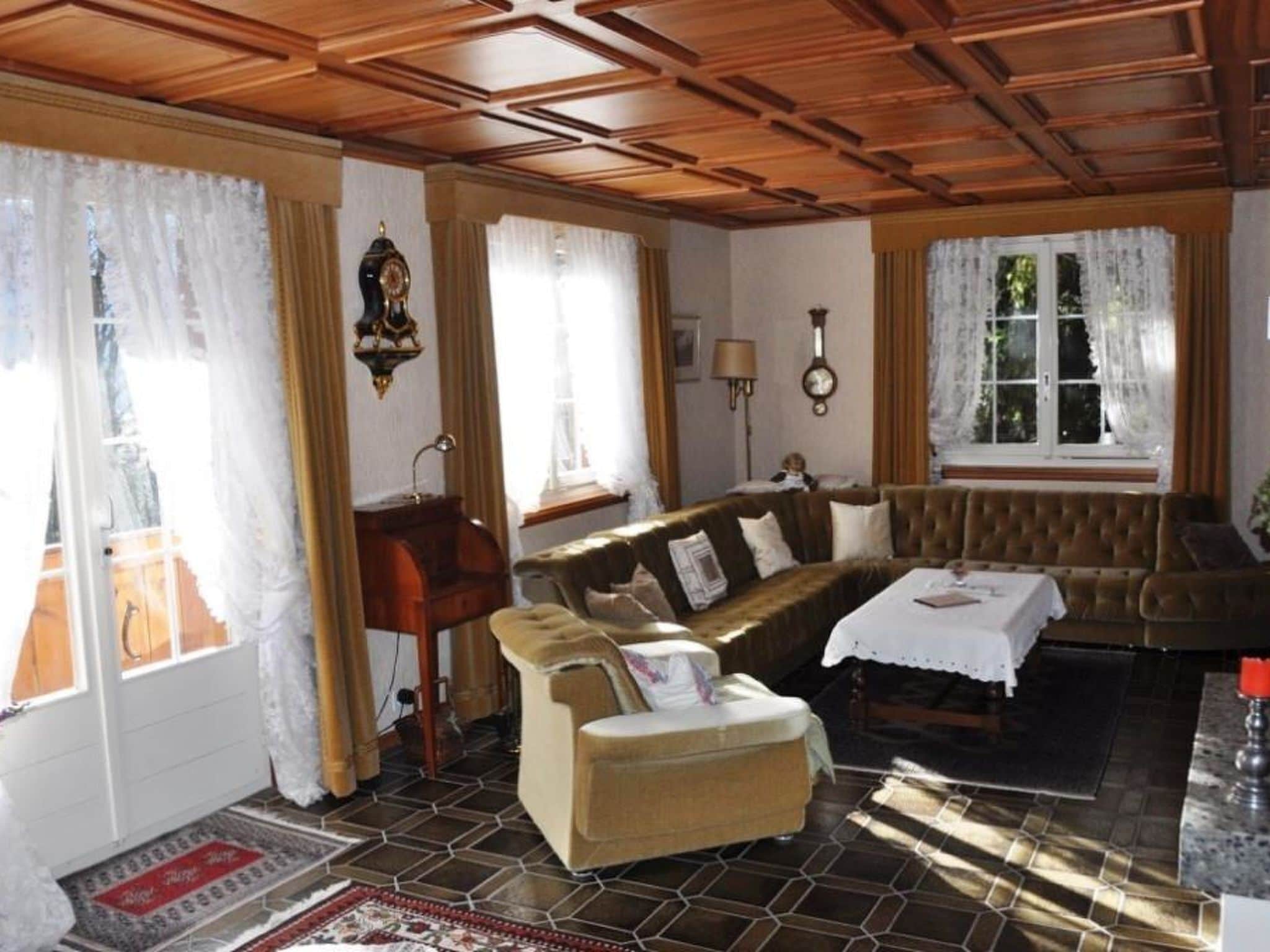 Ein Zimmer mit Holzbalkendecke, einem braunen Sofa, einem weißen Tisch und einem mit Vorhängen drapierten Fenster.