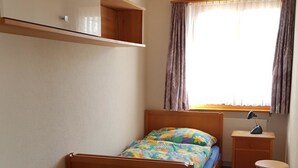 Una habitación pequeña con cama individual, techo de madera, ventana con cortinas, mesita de noche y una silla.