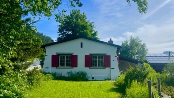 Una casa de un piso con fachada blanca y contraventanas rojas, rodeada de abundante vegetación y un cielo despejado.