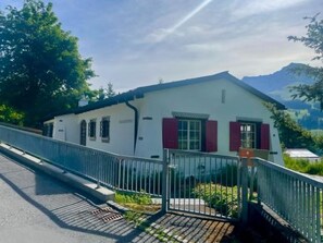 A single-story building with a white facade and red shutters, a metal fence, and a paved entrance. - Baslerdybli Appartement/Fewo, Bad, WC, 3 Schlafräume 4-Zimmerwohnung by Interhome (Adelboden)