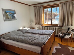 A bedroom with a wooden bed, a window with curtains, a radiator, a chair, and a cowhide rug. - Baslerdybli Appartement/Fewo, Bad, WC, 3 Schlafräume 4-Zimmerwohnung by Interhome (Adelboden)