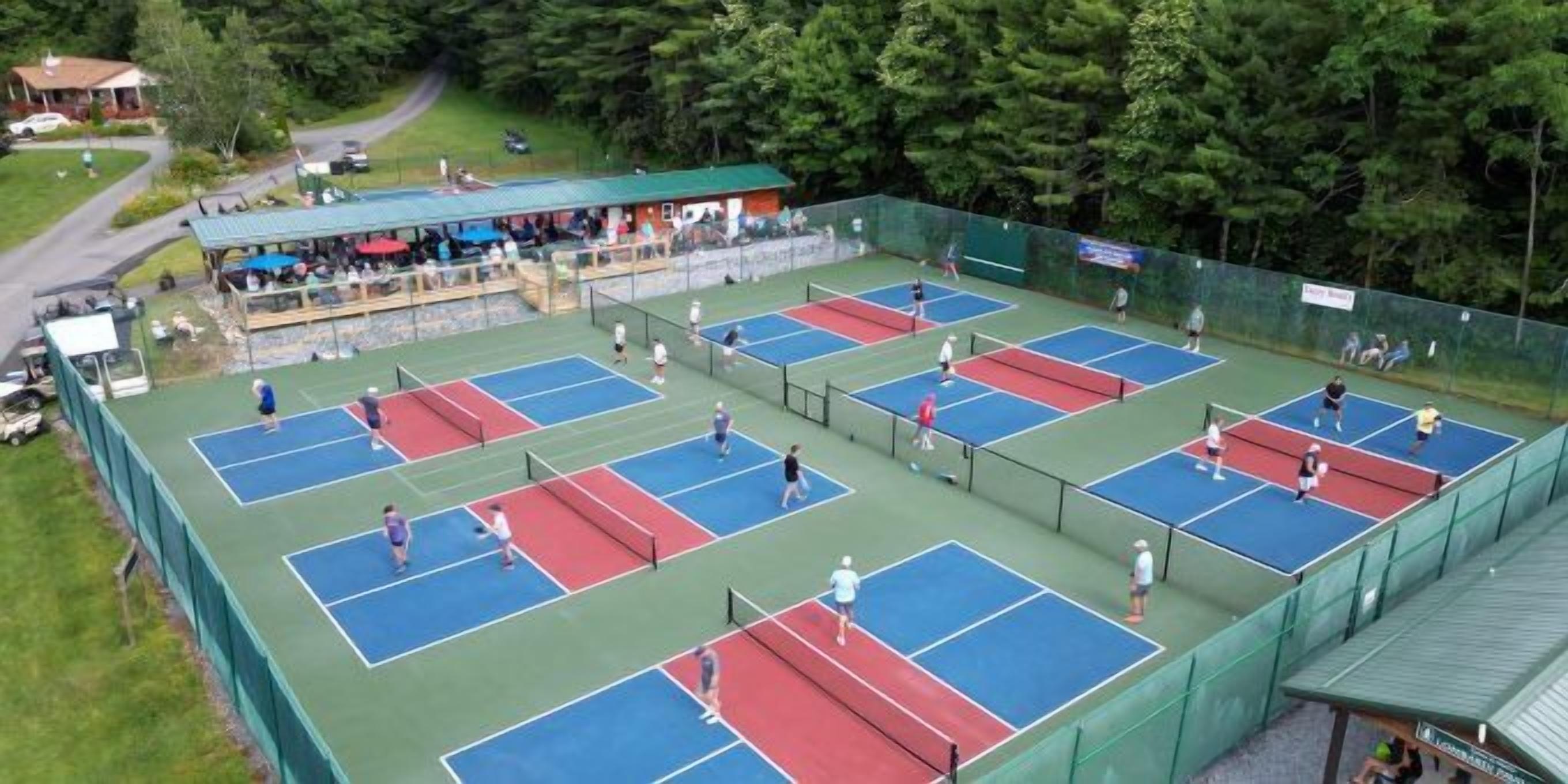 An outdoor tennis court complex with multiple courts, spectator seating, and a covered area.
