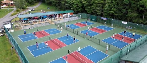 An outdoor tennis court complex with multiple courts, spectator seating, and a covered area.