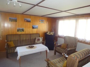 Une pièce avec un mur lambrissé en bois, un canapé, un fauteuil, une petite table recouverte d’une nappe blanche et une fenêtre avec des rideaux.