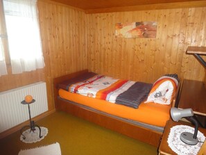 Een kleine kamer met een enkel bed, houten wanden, een radiator, een bureau met lamp en een raam met gordijnen.