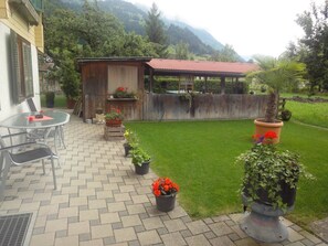 Eine Terrasse mit Tisch und Stühlen, eine Holzhütte mit rotem Dach und ein gepflegter Rasen.