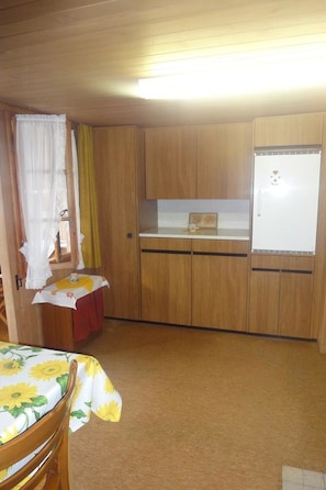 Eine kleine Küche mit Holzausstattung, einem Kühlschrank, einem Tisch mit Stoffauflage und einem Stuhl mit Blumenmuster.