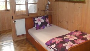Un llit individual amb una edredó floral, un radiador, una finestra amb cortines de galeria i un quadre a la paret, així com també un lluminari al sostre.