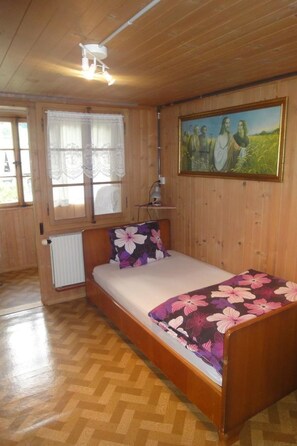 Ein Einzelbett mit Blumenmuster, ein Heizkörper, ein Fenster mit Spitzenvorhängen, ein Bild an der Wand und eine Deckenleuchte.