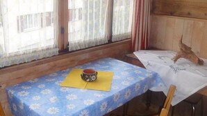 Una habitació de fusta amb una taula coberta amb un mantell blau amb flors, dues cadires de fusta i una finestra amb cortines translúcides.