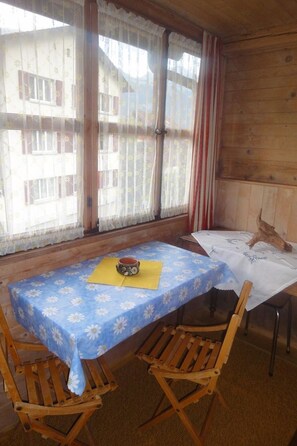 Ein Zimmer mit Holzboden, einem Tisch mit blühendem, blauem Tischtuch, zwei Holzstühlen und einem Fenster mit durchsichtigen Vorhängen.