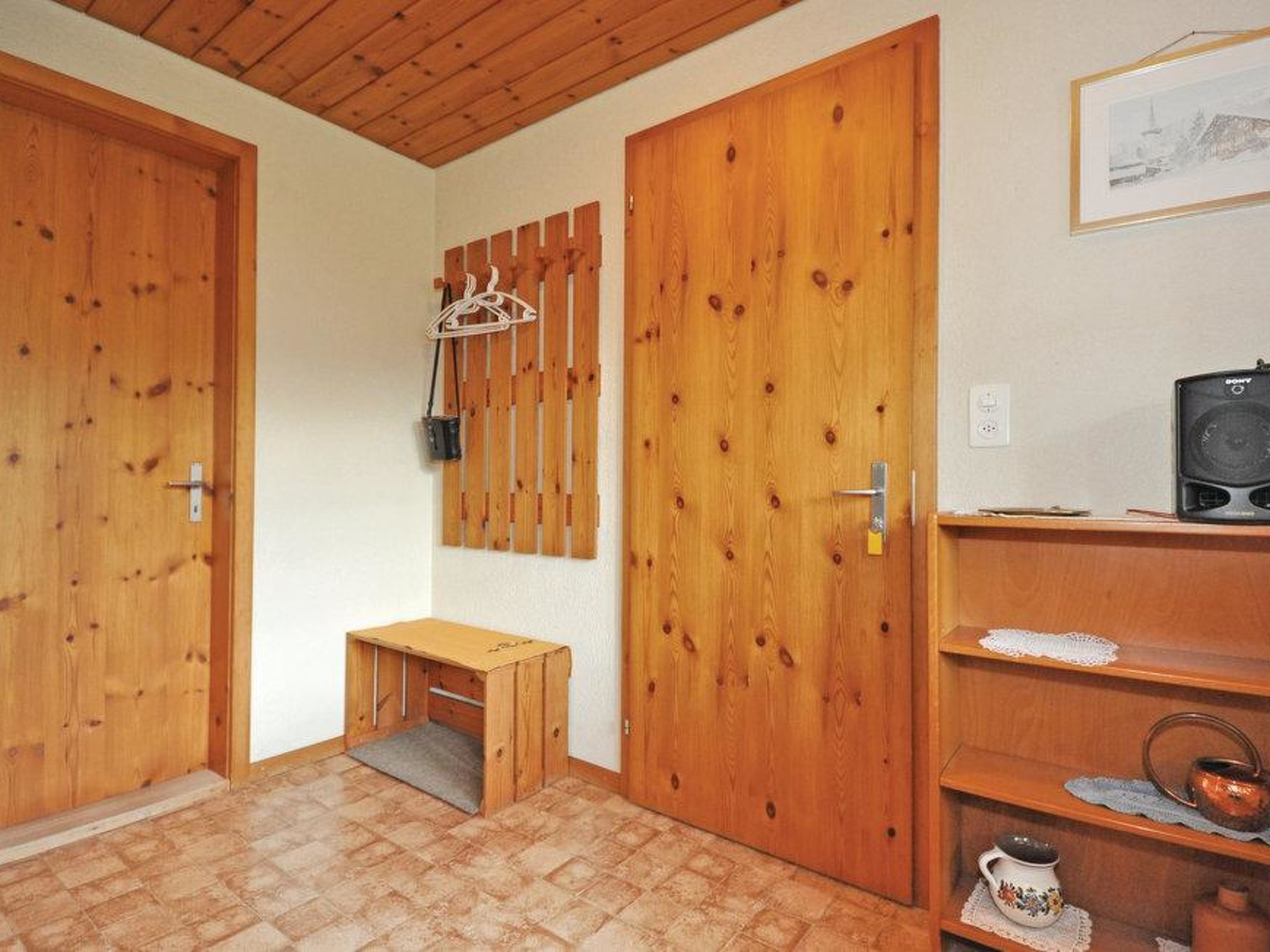 Une pièce avec des portes en bois, un petit banc en bois, une étagère contenant des ustensiles de cuisine et un tableau encadré au mur.