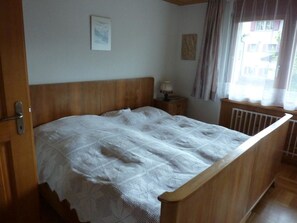A bedroom with a wooden bed, a nightstand, a lamp, and a window with curtains. - Abesunne Appartement/Fewo, Dusche, WC, 1 Schlafraum Sarbach - Erdgeschoss by Interhome (Frutigen)