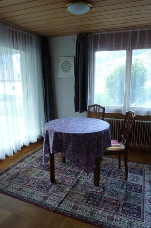 A room with a round table covered by a patterned tablecloth, a chair, a radiator, and a window with curtains. - Abesunne Appartement/Fewo, Dusche, WC, 1 Schlafraum Sarbach - Erdgeschoss by Interhome (Frutigen)