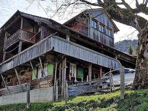 A two-story wooden house with a balcony, a car parked in front, and a tree to the right.