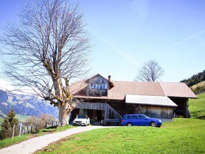 Ein Holzhaus mit rotem Dach, ein geparktes Auto davor und ein Baum im Vordergrund.