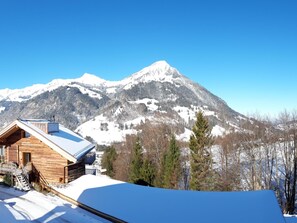 Eine Holzhütte mit schneebedecktem Dach vor einer Berglandschaft mit Schnee bedeutenen Gipfeln, vor blauem, wolkenlosem Himmel.