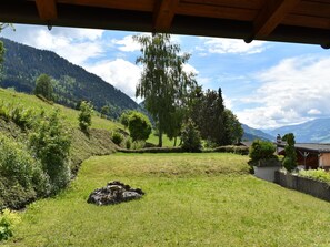 Ein grasbewachsenes Freigelände mit einer hölzernen Überdachung, einem großen Baum und einer Berglandschaft im Hintergrund.